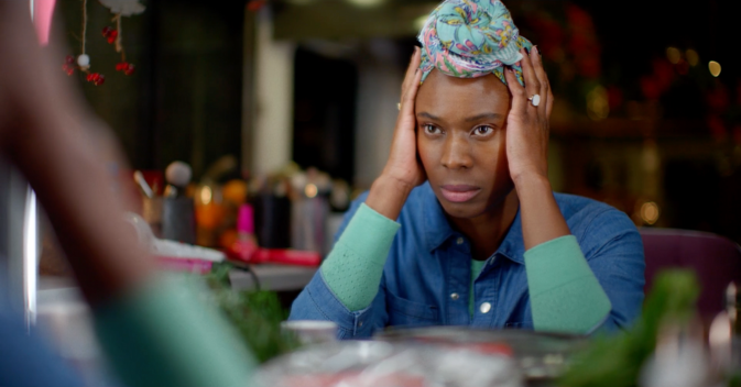 A make up up artist is sitting in a TV production trailer. She has her head in her hands and is looking at herself in the mirror with an glum expression.